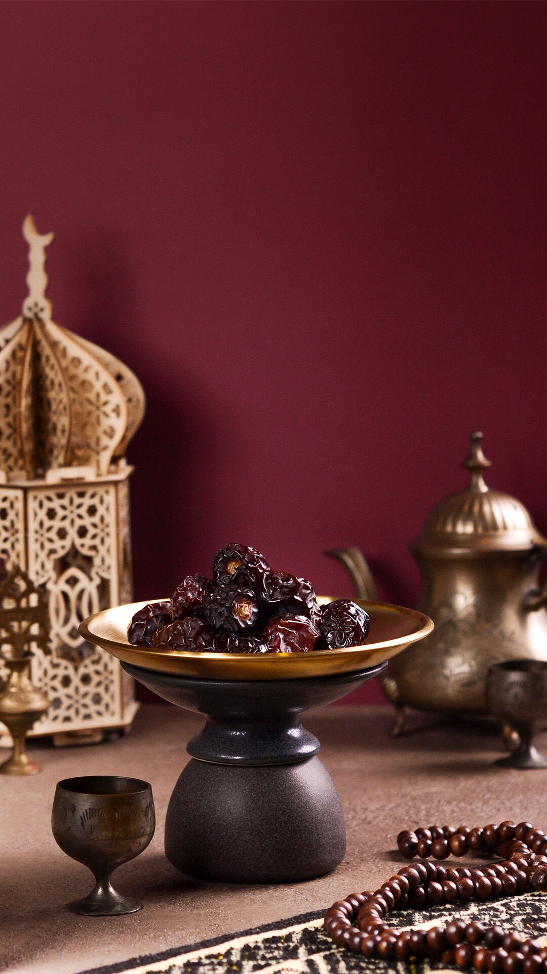Bowl of dates with traditional Arabic coffee set, lantern, and prayer beads on a Ramadan-themed table against a rich burgundy backdrop.
